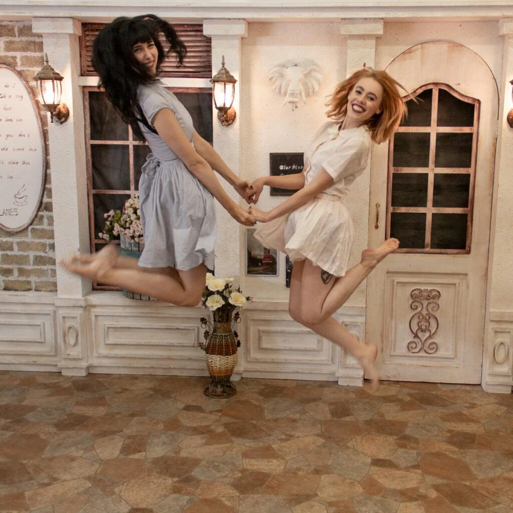 Two women jumping and smiling indoors in stylish dresses, exuding happiness.